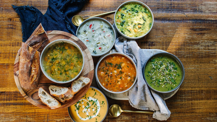 Assorted bowls of soup on a wooden table with bread and spoons.